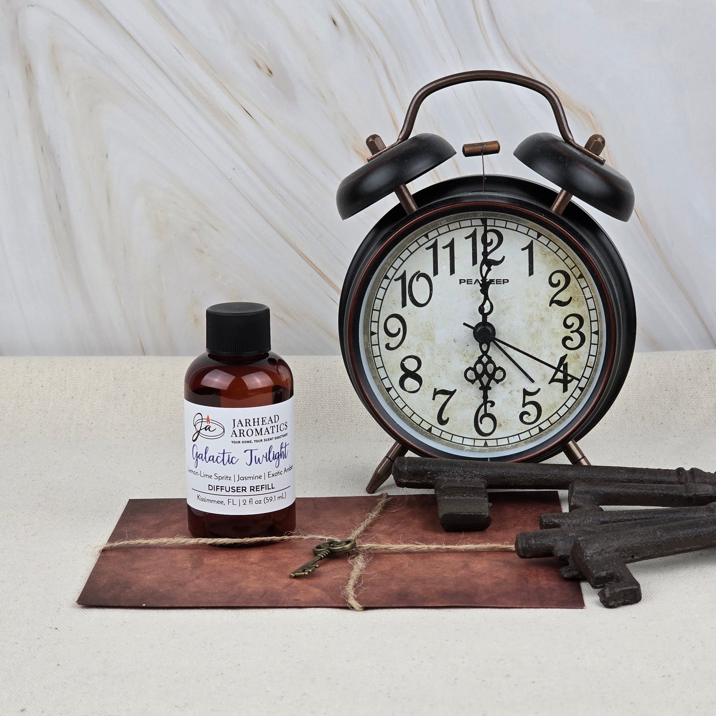 A vintage alarm clock, an old-fashioned key, two large metal keys, and Jarhead Aromatics' Galactic Twilight Diffuser Refill—Amber, Jasmine & Sandalwood—rest on a tied brown envelope set against a light marbled background.