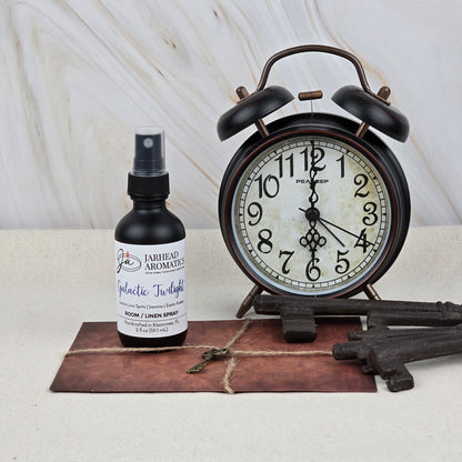 A Jarhead Aromatics Galactic Twilight Room & Linen Spray (Amber, Bergamot & Sandalwood), a vintage alarm clock, a tied old envelope, a small key, and two large antique keys are arranged on beige for an inviting home fragrance scene.