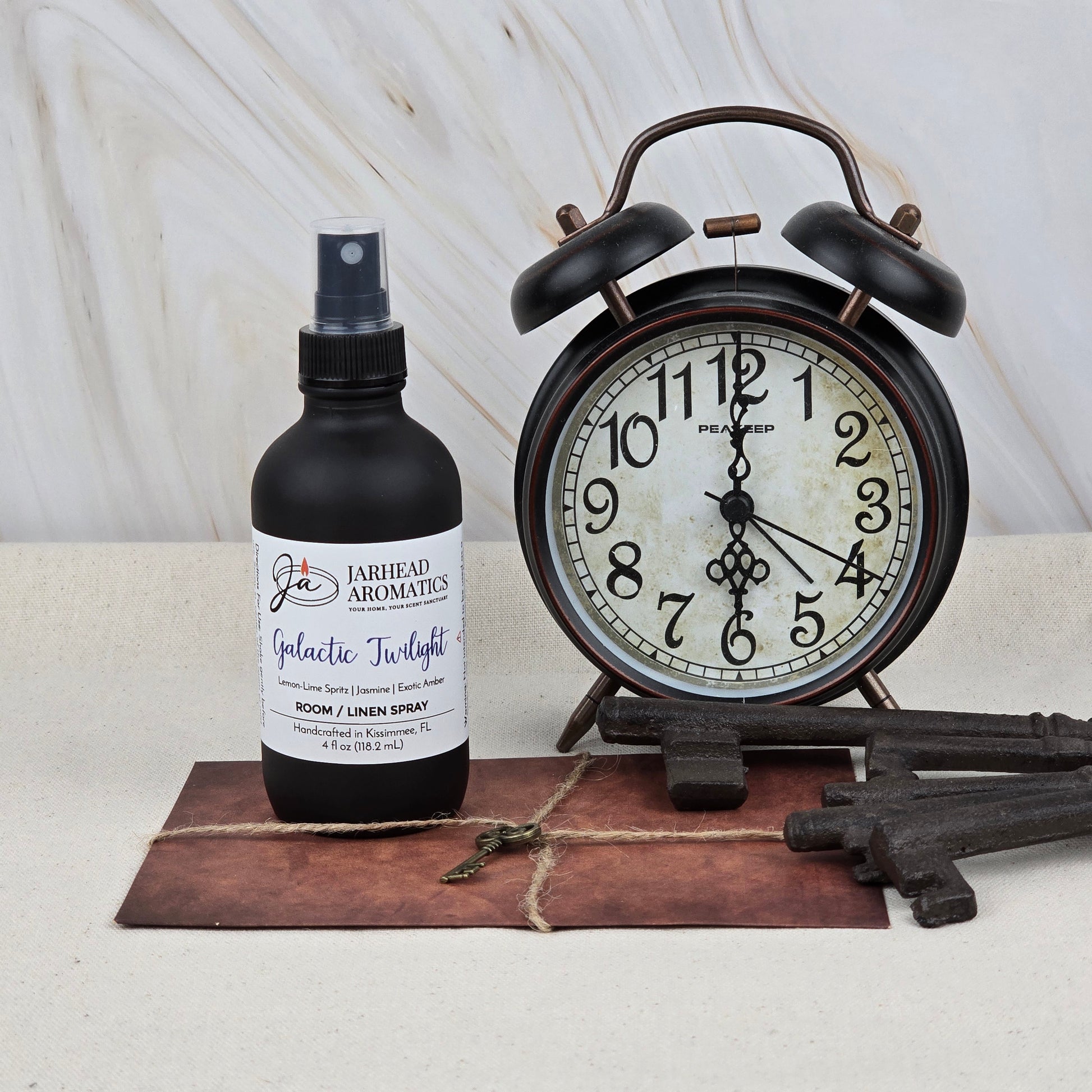 A black vintage alarm clock, three decorative keys, and Jarhead Aromatics’ Galactic Twilight Room & Linen Spray—featuring amber, bergamot, and sandalwood—sit on a brown envelope with a metal key against a light marble background.