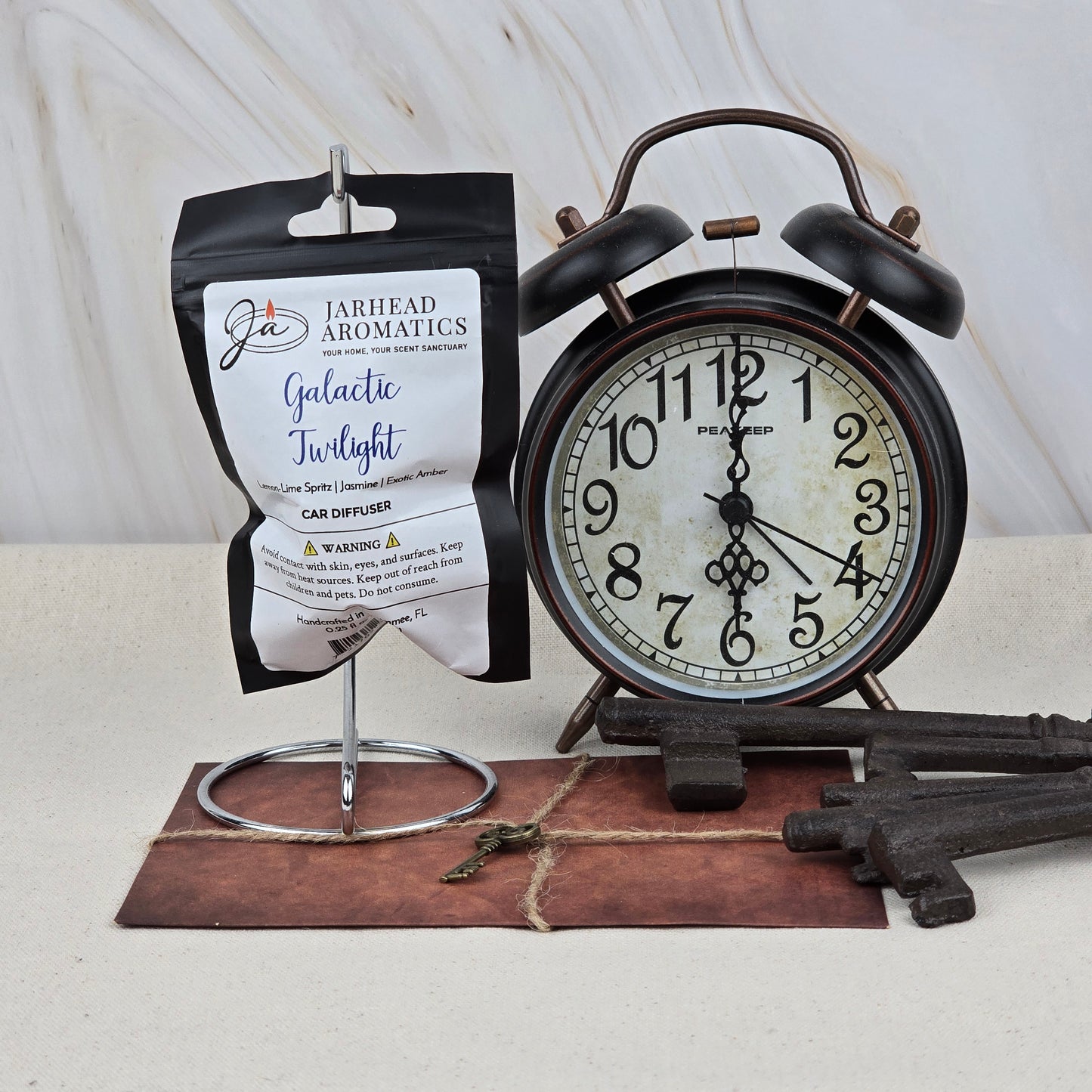 A black alarm clock, three vintage keys, and the Jarhead Aromatics Galactic Twilight Car Diffuser | Amber, Citrus & Sandalwood are arranged on a surface with a marble backdrop. A small key rests on a brown notepad beneath the clock.