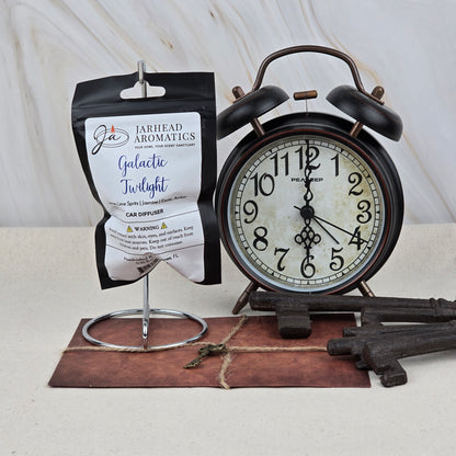 A black alarm clock, three vintage keys, and the Jarhead Aromatics Galactic Twilight Car Diffuser | Amber, Citrus & Sandalwood are arranged on a surface with a marble backdrop. A small key rests on a brown notepad beneath the clock.