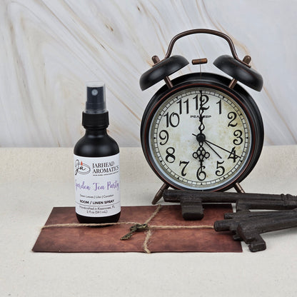 A black alarm clock, Garden Tea Party Room & Linen Spray by Jarhead Aromatics, three vintage keys, and an old envelope with a small key on top are arranged on a light surface against a marble background.