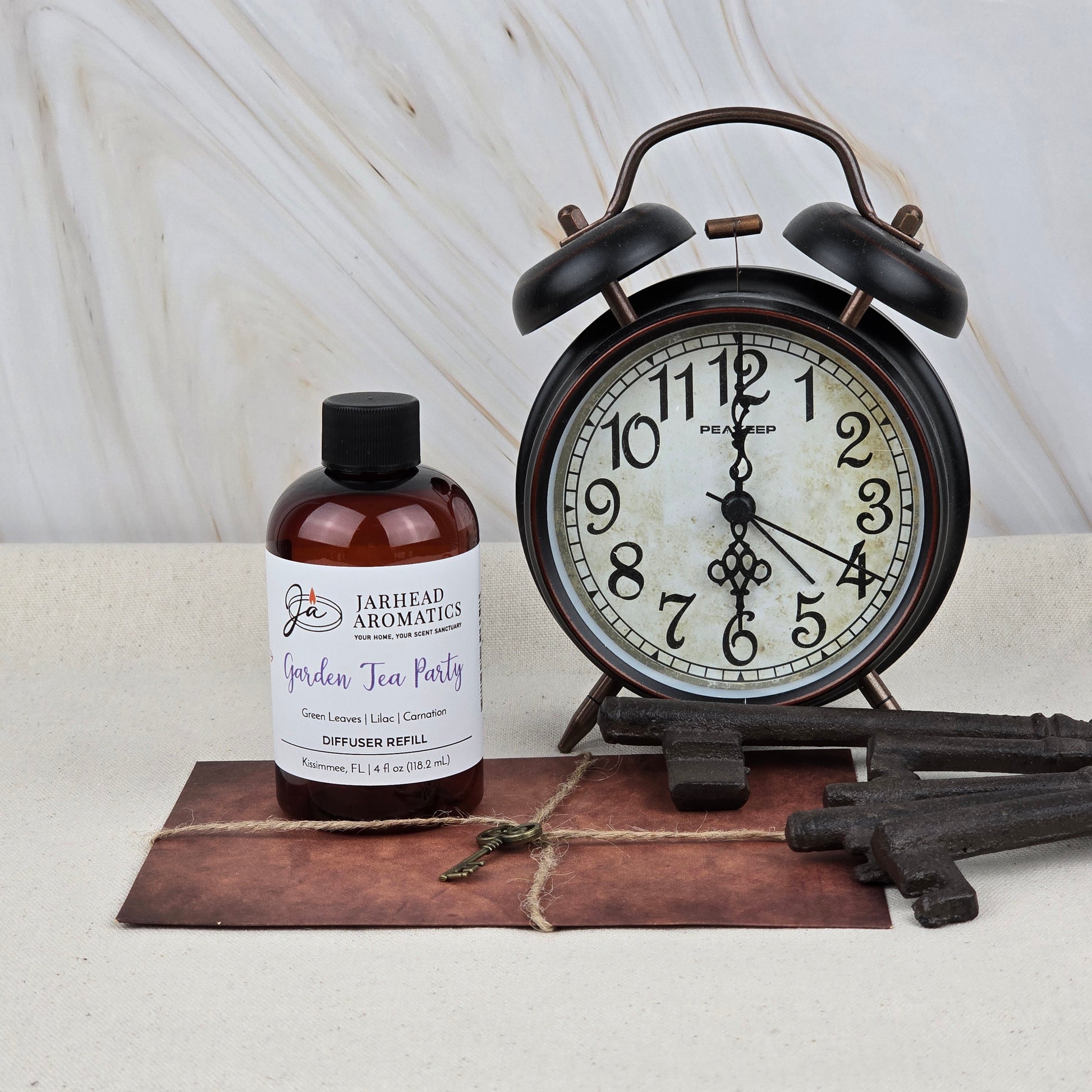 A brown bottle of Jarhead Aromatics’ Garden Tea Party Diffuser Refill (Rose, Jasmine & Vanilla) sits by a vintage black alarm clock, ornate keys, and a tied envelope on marble, capturing the charm of lush garden scents.