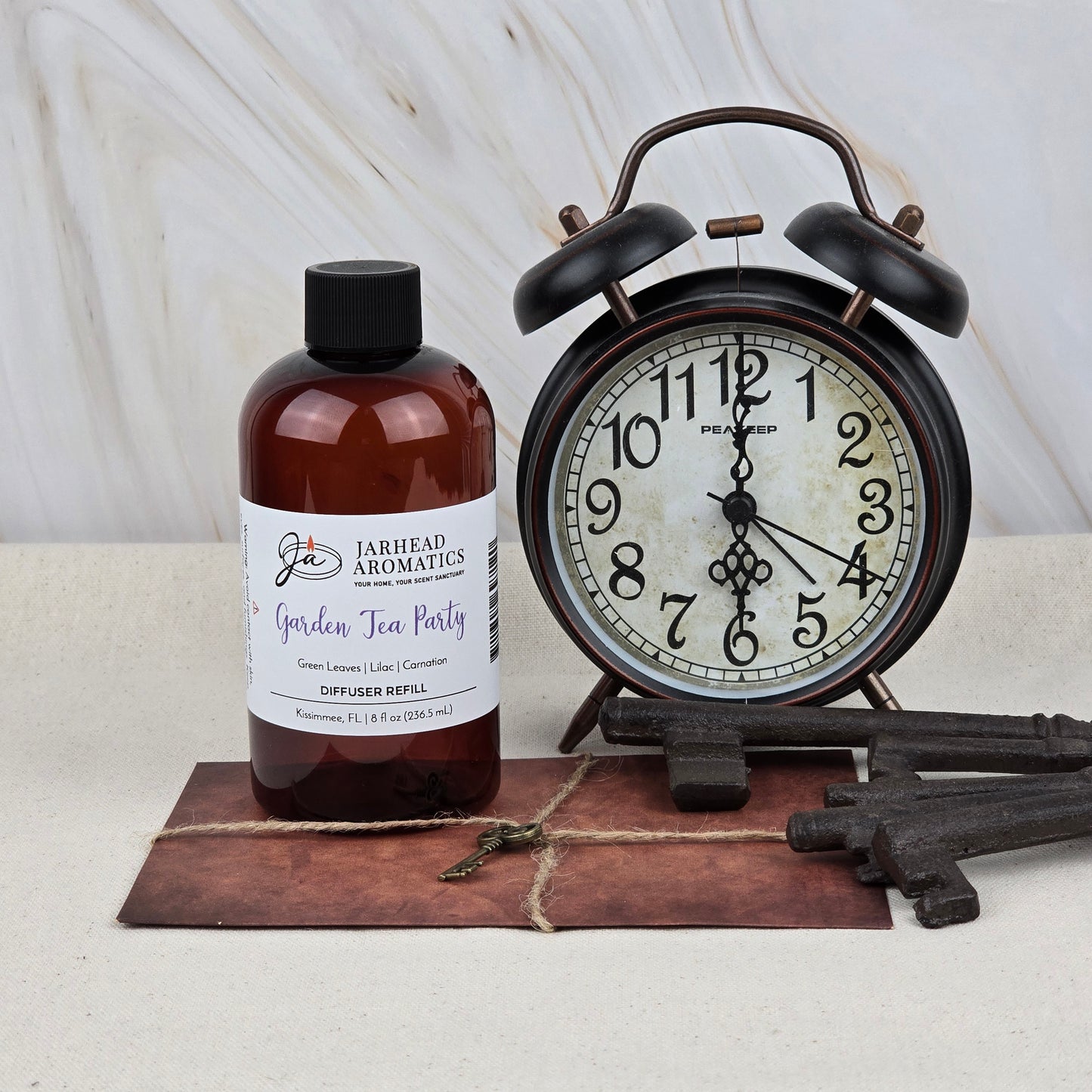A bottle of Jarhead Aromatics Garden Tea Party Diffuser Refill | Rose, Jasmine & Vanilla sits on a tile next to a vintage black alarm clock and old-fashioned keys, evoking a charming scene that hints at its floral garden fragrance.