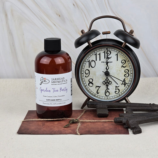 A bottle of Jarhead Aromatics Garden Tea Party Diffuser Refill | Rose, Jasmine & Vanilla sits on a tile next to a vintage black alarm clock and old-fashioned keys, evoking a charming scene that hints at its floral garden fragrance.