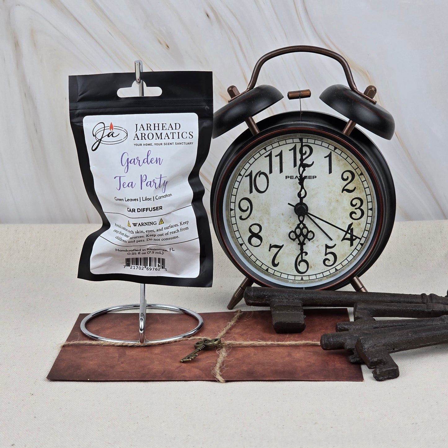 A packet of Jarhead Aromatics Garden Tea Party Car Diffuser, an elegant floral and garden-inspired air freshener, hangs beside a vintage alarm clock, decorative keys, and a leather-bound book on a light-colored surface.