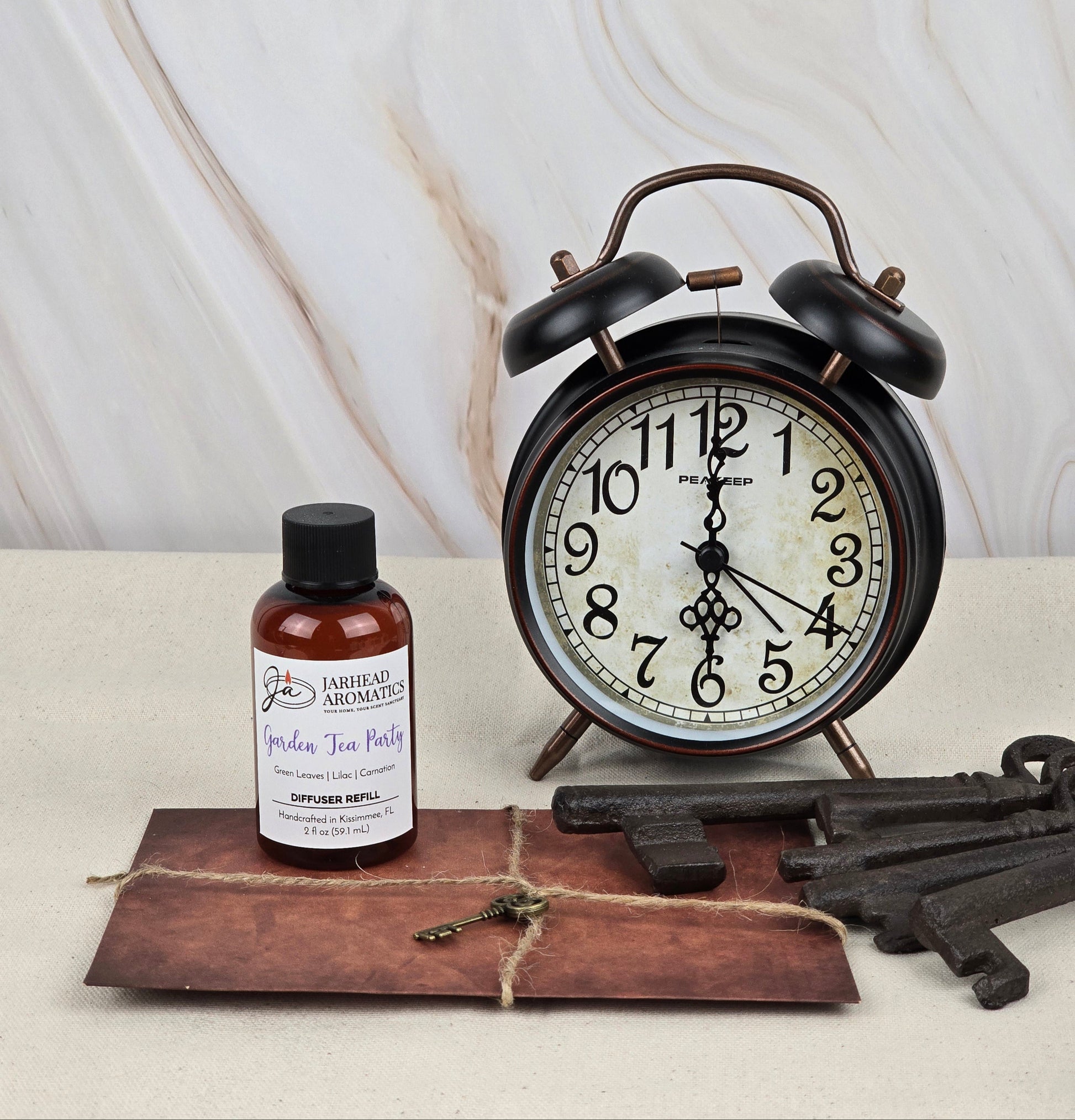 A vintage-style alarm clock with black bells sits on a table. Nearby are a dark brown envelope tied with string, a bottle of Jarhead Aromatics Garden Tea Party Reed Diffuser Refill exuding floral scents, and metal keys, all against a marbled background.