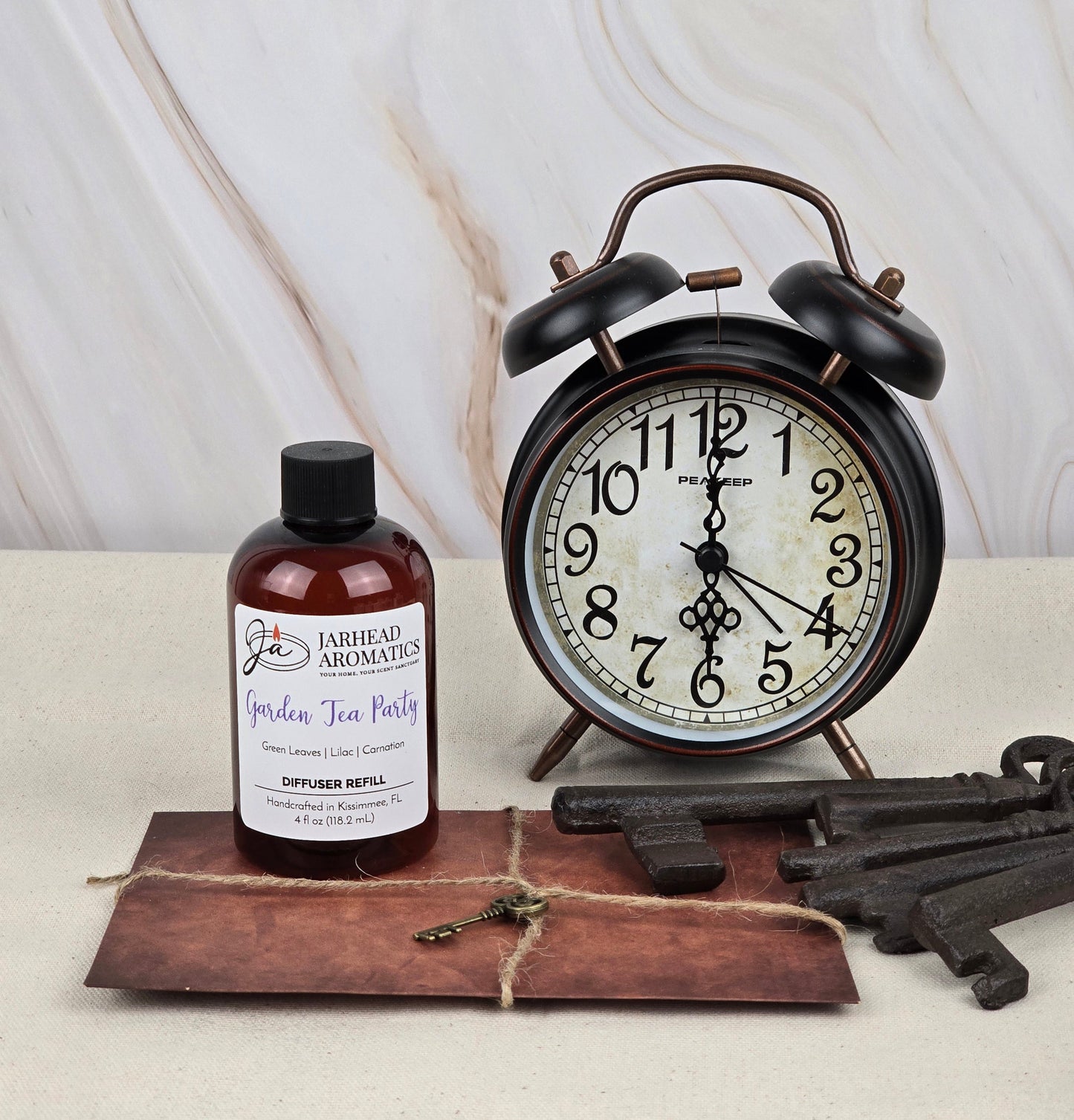 A vintage-style alarm clock, large rustic keys, a brown envelope, and a bottle labeled Garden Tea Party - Reed Diffuser Refill from Jarhead Aromatics with floral scents are arranged on a light surface against a marble-like background.