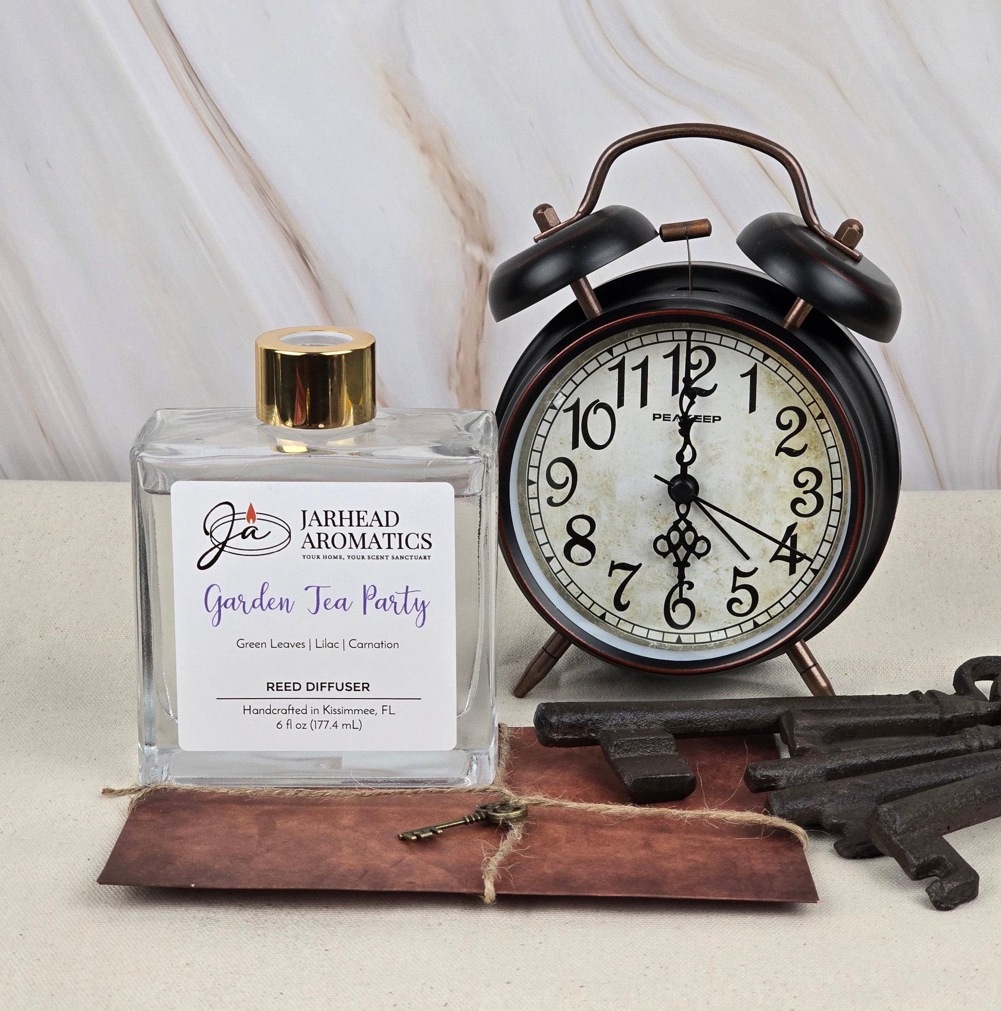 A vintage alarm clock is beside a glass bottle with a gold cap labeled Jarhead Aromatics Garden Tea Party - Reed Diffuser, emitting floral aromas. Nearby, three rustic keys rest on a leather pad atop a marble-like surface.