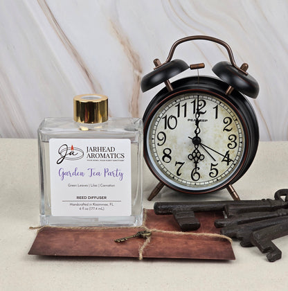 A vintage alarm clock is beside a glass bottle with a gold cap labeled Jarhead Aromatics Garden Tea Party - Reed Diffuser, emitting floral aromas. Nearby, three rustic keys rest on a leather pad atop a marble-like surface.
