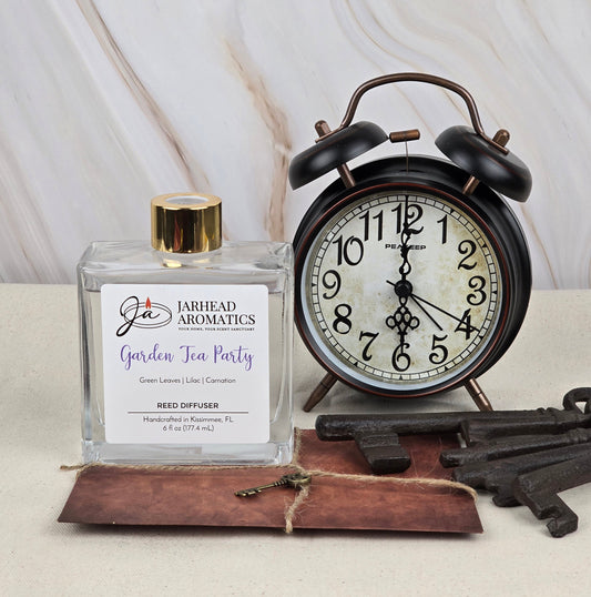 A vintage alarm clock is beside a glass bottle with a gold cap labeled Jarhead Aromatics Garden Tea Party - Reed Diffuser, emitting floral aromas. Nearby, three rustic keys rest on a leather pad atop a marble-like surface.