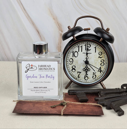 A Garden Tea Party reed diffuser by Jarhead Aromatics with floral scents sits beside a vintage-style black alarm clock showing 10:09. Below, a brown leather-bound notebook and three antique keys rest elegantly on a marble background.