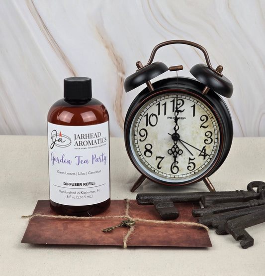 A bottle of Jarhead Aromatics Garden Tea Party - Reed Diffuser Refill, exuding rich floral scents, is placed beside a vintage-style alarm clock and three old-fashioned keys on a brown envelope against a marbled background.