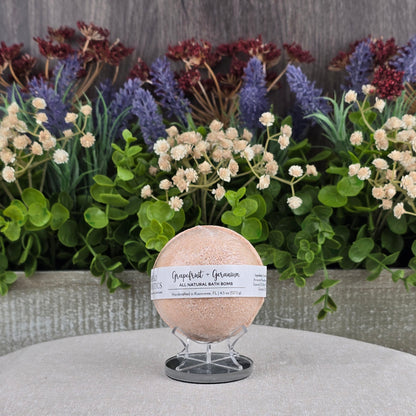 The Grapefruit + Geranium Bath Bomb - All Natural from Jarhead Aromatics rests on light gray fabric with green leaves and colorful flowers behind it, highlighting its natural ingredients.