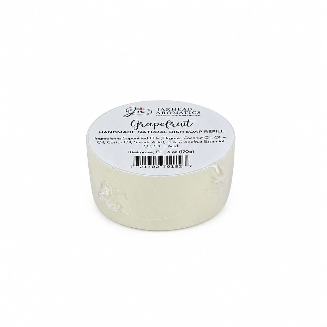 A round, white bar labeled “Grapefruit Handmade Natural Solid Dish Soap | Citrus Kitchen Cleaning Bar with Grapefruit Essential Oil” from Jarhead Aromatics rests on a white surface. The handmade natural dish soap displays its ingredients and barcode on the label.