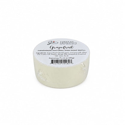 A round, white bar labeled “Grapefruit Handmade Natural Solid Dish Soap | Citrus Kitchen Cleaning Bar with Grapefruit Essential Oil” from Jarhead Aromatics rests on a white surface. The handmade natural dish soap displays its ingredients and barcode on the label.