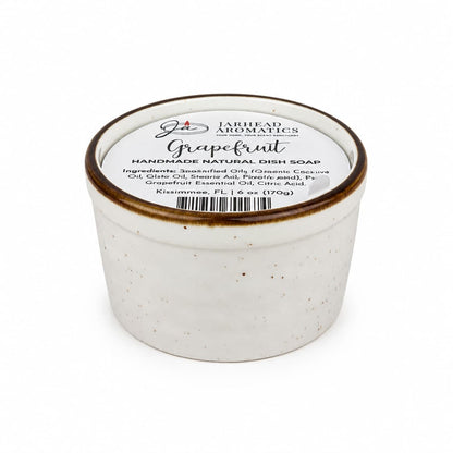 A white ceramic jar of Jarhead Aromatics Grapefruit Handmade Natural Solid Dish Soap | Citrus Kitchen Cleaning Bar with Grapefruit Essential Oil is photographed on a white background, its lid displaying an ingredient list. This plant-based, eco-friendly dish soap offers natural cleaning power paired with the invigorating aroma of grapefruit—an essential for any green kitchen.
