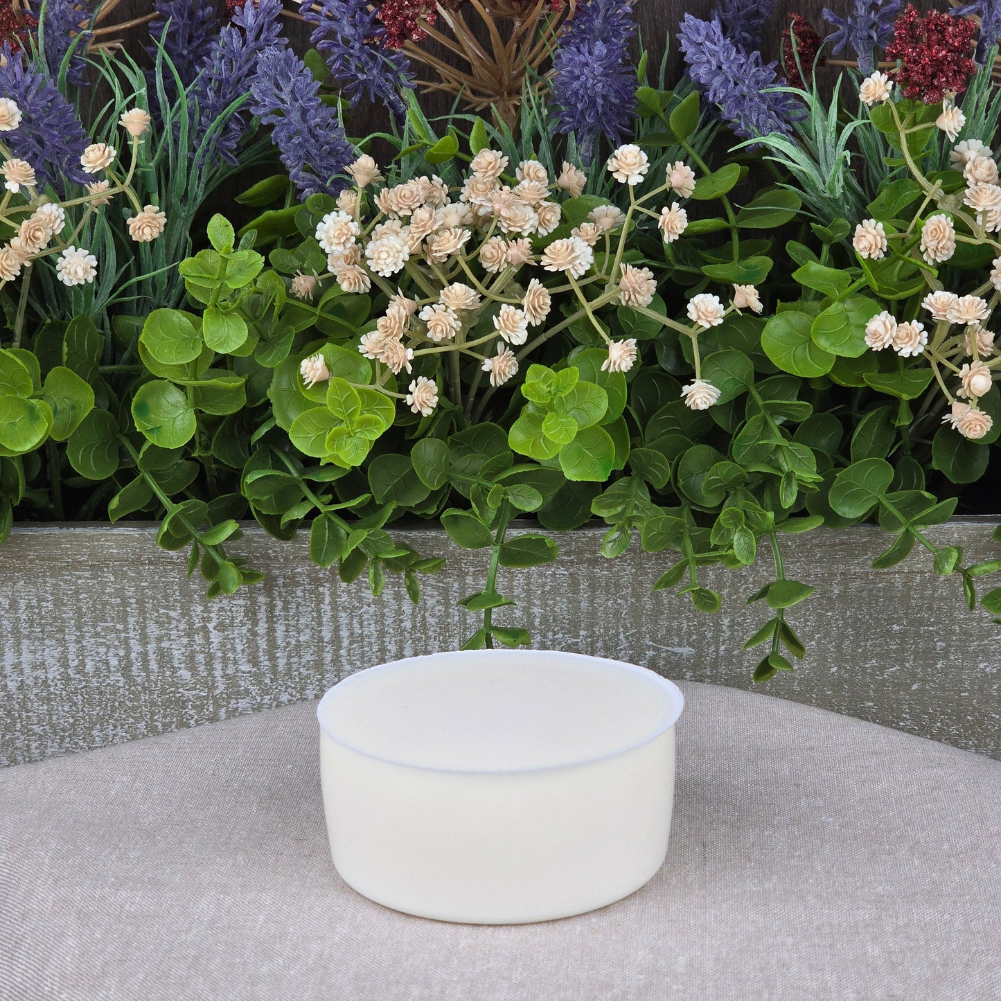 A round, white Lemon Handmade Natural Solid Dish Soap by Jarhead Aromatics rests on a beige surface in front of a planter with green leaves, white flowers, and purple lavender.