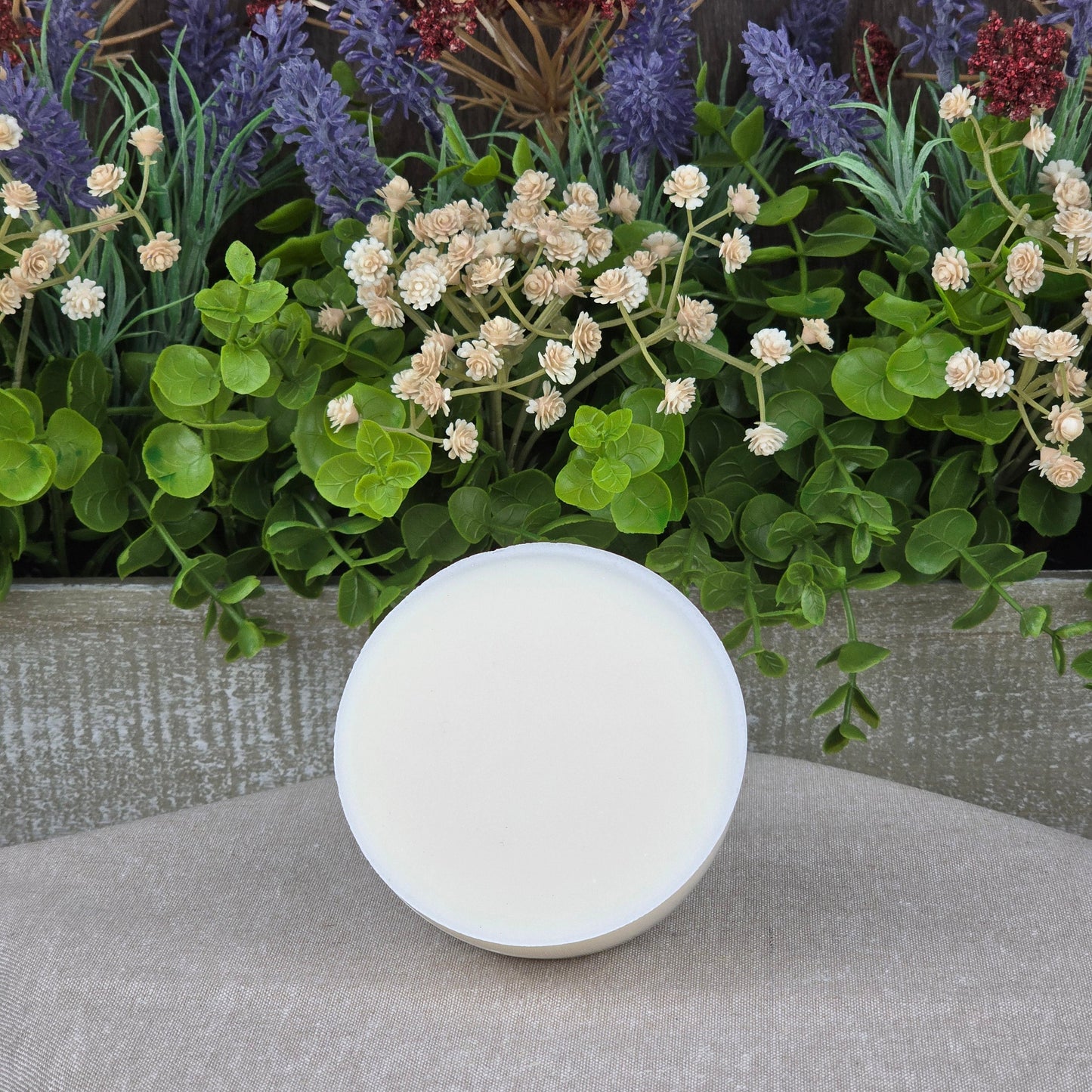 A round, white container of Jarhead Aromatics Lemon Handmade Natural Solid Dish Soap with a closed lid sits on a beige surface, in front of a wooden box filled with green foliage, white flowers, and purple lavender.