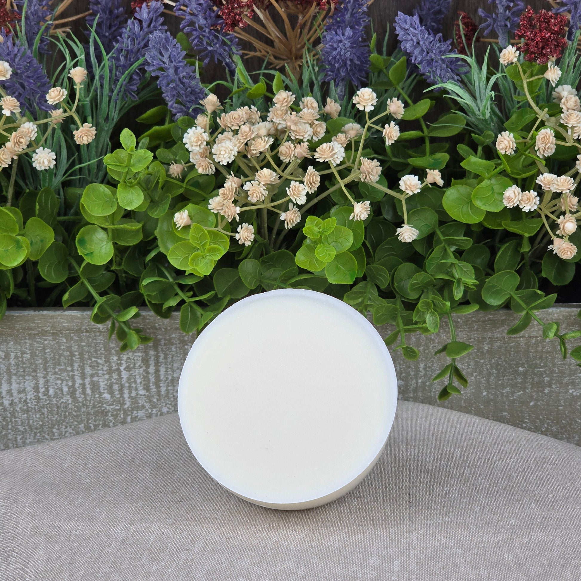 A round, white container of Jarhead Aromatics Lemon Handmade Natural Solid Dish Soap with a closed lid sits on a beige surface, in front of a wooden box filled with green foliage, white flowers, and purple lavender.