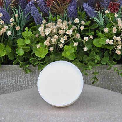 A round, white container of Jarhead Aromatics Lemon Handmade Natural Solid Dish Soap with a closed lid sits on a beige surface, in front of a wooden box filled with green foliage, white flowers, and purple lavender.