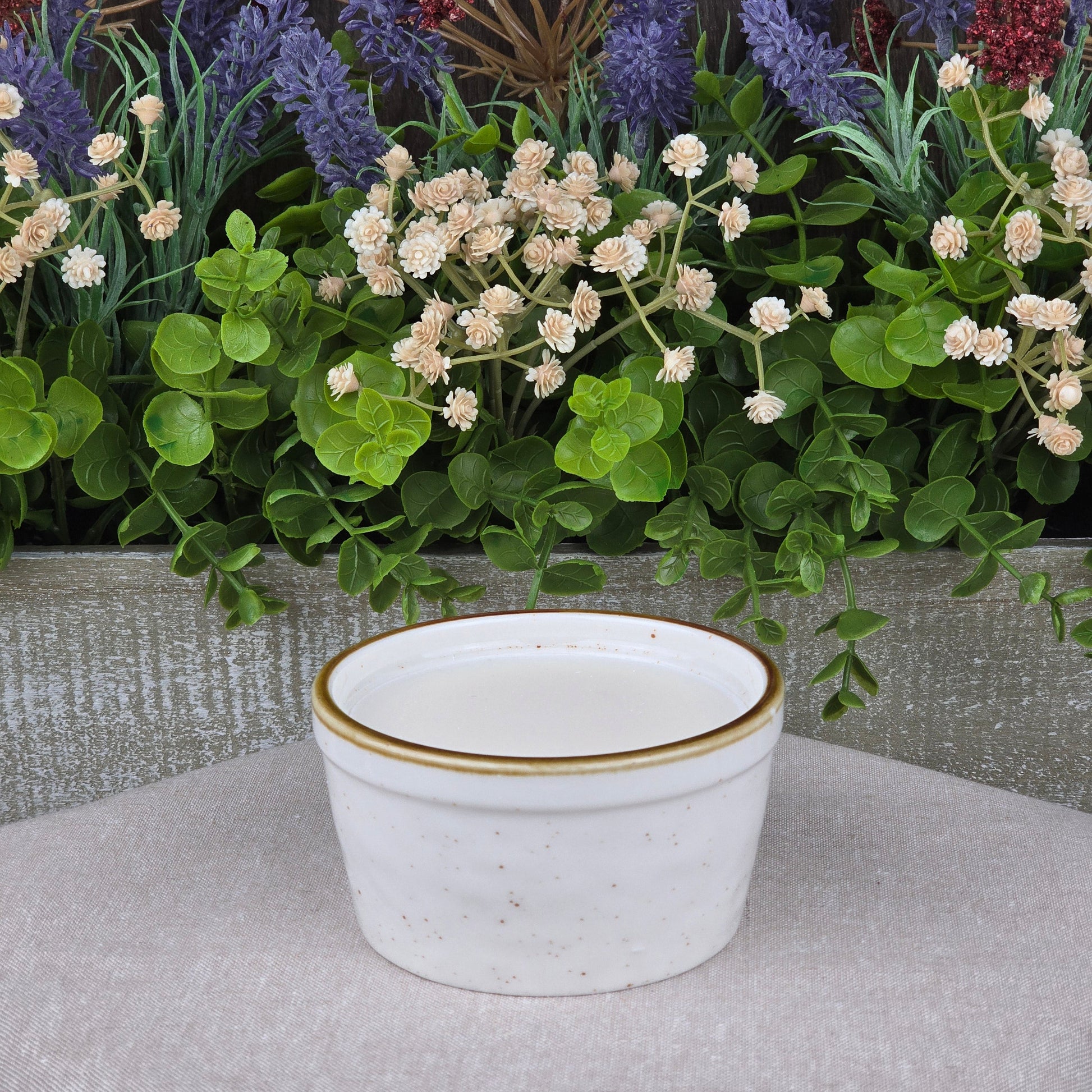 A small, white ceramic bowl with a thin gold rim holds Jarhead Aromatics’ Lemon Handmade Natural Solid Dish Soap on a beige surface, set before green leaves and white and purple flowers.