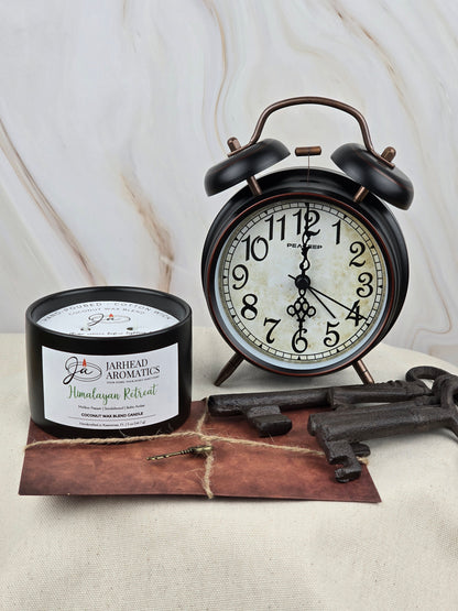 A Himalayan Retreat Coconut Wax Blend Candle by Jarhead Aromatics, featuring cedar, sandalwood, and amber, rests next to a vintage alarm clock on a textured surface, with three old keys in front.