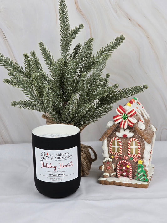 A black Holiday Hearth - Soy Wax Candle by Jarhead Aromatics sits next to a candy-decorated gingerbread house, framed by a frosted evergreen branch, emitting a warm festive blend of aromas.