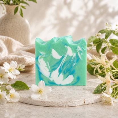 A green and blue marbled bar of Jarhead Aromatics Honeysuckle + Jasmine Handmade Coconut Milk Artisan Soap, known for its fresh floral garden scent, rests on a round stone surface, surrounded by white flowers, green leaves, and a beige textured cloth in a softly lit, natural setting.