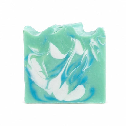A rectangular bar of Jarhead Aromatics’ Honeysuckle + Jasmine Handmade Coconut Milk Artisan Soap features a green base with blue and white swirling patterns and a wavy, uneven top, creating a fresh floral garden look.