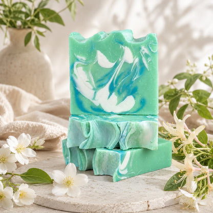 Three bars of Jarhead Aromatics’ Honeysuckle + Jasmine Handmade Coconut Milk Artisan Soap, featuring a marbled blend of green and white, are stacked on a stone surface amidst delicate white flowers and fresh greenery. In the softly blurred background, a beige vase and gentle fabric add to the inviting, botanical ambiance.