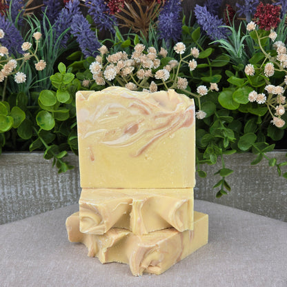 Three IPA + Pine Handmade Craft Beer Artisan Soaps by Jarhead Aromatics, featuring creamy yellow swirls and textured tops, are stacked in front of green leaves and colorful flowers on a neutral fabric background.