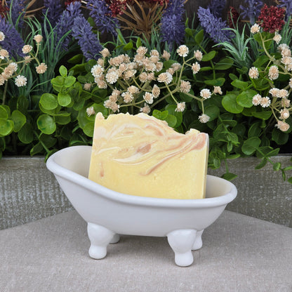 The IPA + Pine Handmade Craft Beer Artisan Soap by Jarhead Aromatics rests in a small, white, bathtub-shaped dish with green and purple artificial flowers in the background.