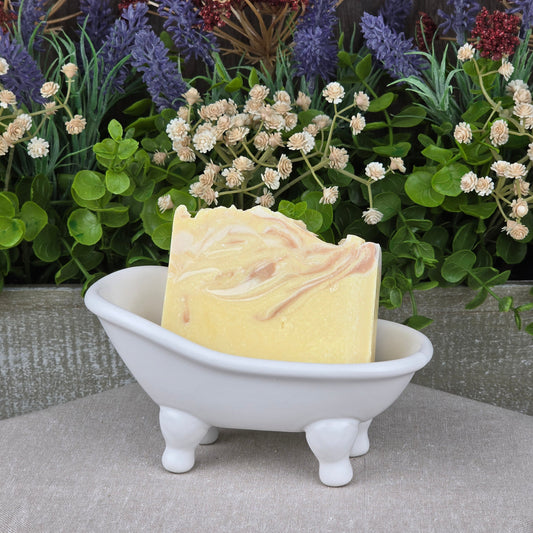The IPA + Pine Handmade Craft Beer Artisan Soap by Jarhead Aromatics rests in a small, white, bathtub-shaped dish with green and purple artificial flowers in the background.