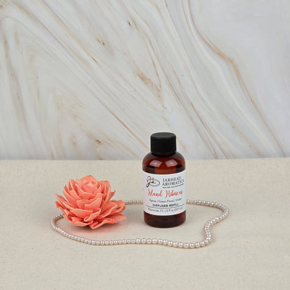 A small amber bottle of Jarhead Aromatics Island Hibiscus Diffuser Refill, featuring tropical melon, lime, and floral notes, sits on a beige surface with a pink artificial flower and pearls against a light marbled background.