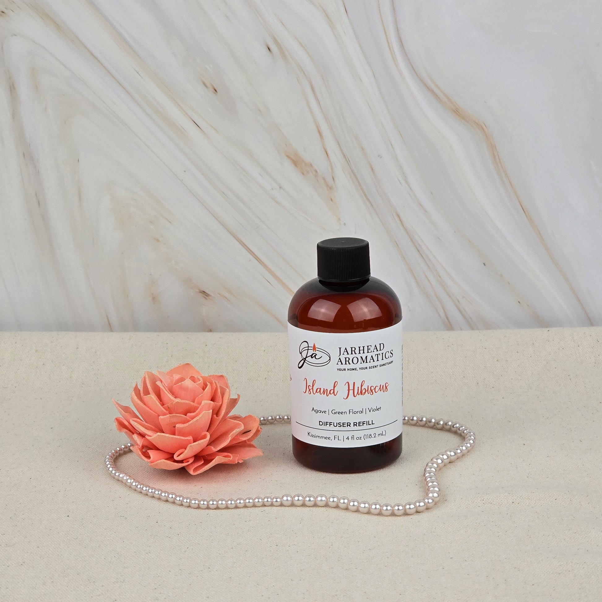 A bottle of Jarhead Aromatics' Island Hibiscus Diffuser Refill | Tropical Melon, Lime & Floral Home Fragrance sits on a beige surface beside a pink faux flower and pearls, adding a tropical scent to the marble-patterned background.