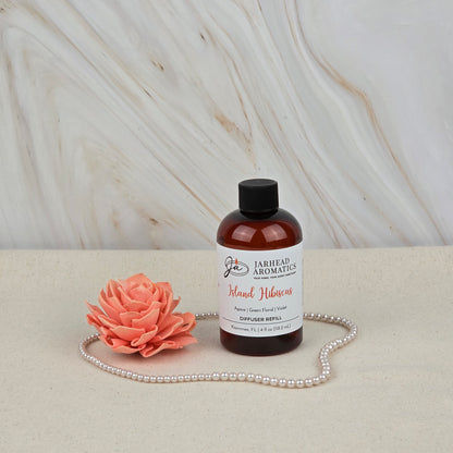 A bottle of Jarhead Aromatics' Island Hibiscus Diffuser Refill | Tropical Melon, Lime & Floral Home Fragrance sits on a beige surface beside a pink faux flower and pearls, adding a tropical scent to the marble-patterned background.