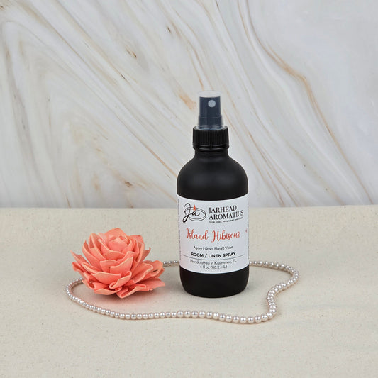 A black Jarhead Aromatics Island Hibiscus Room & Linen Spray rests on a light surface with a string of pearls and a peach artificial flower, set against marble décor, highlighting this tropical floral home fragrance spray.