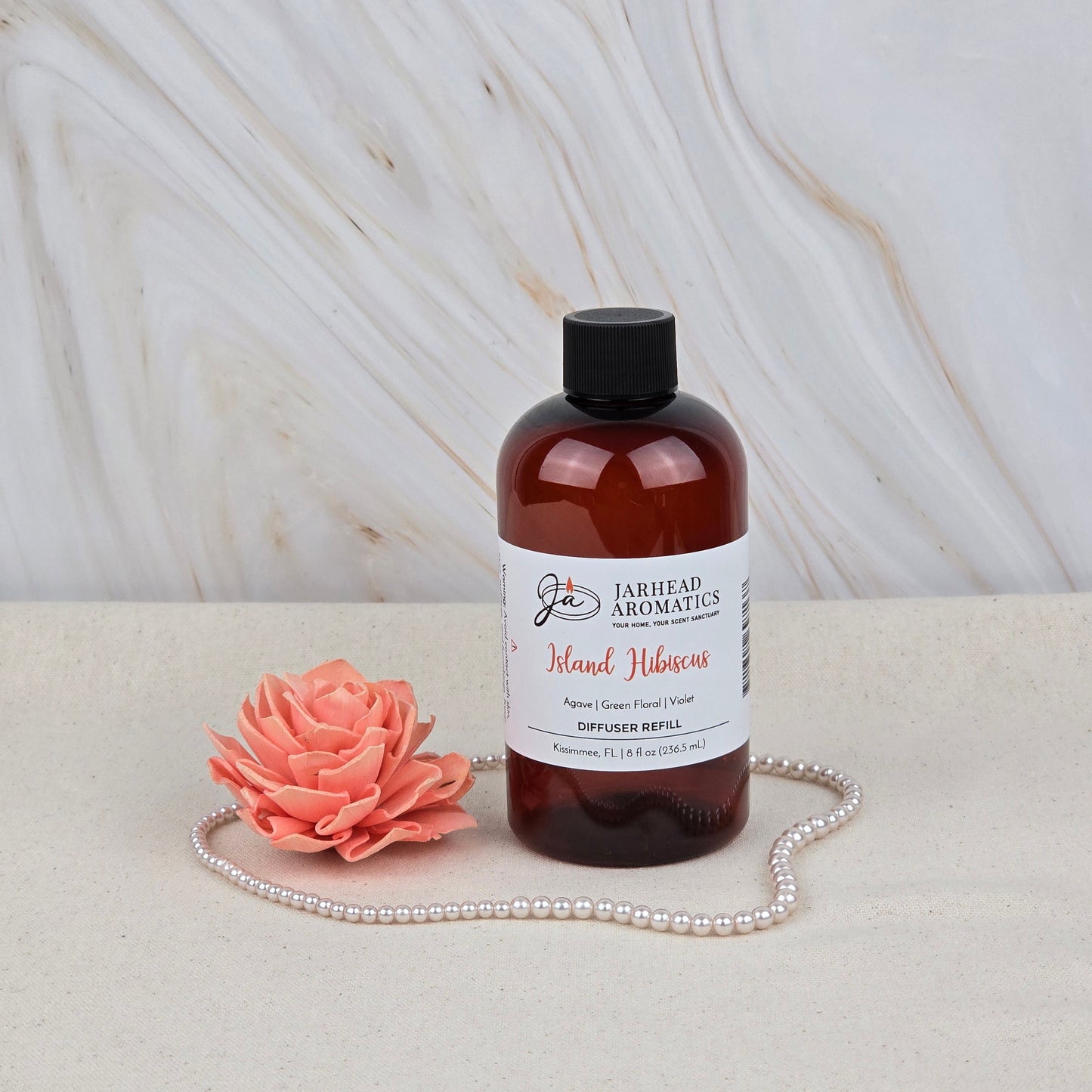 A brown Jarhead Aromatics Island Hibiscus Diffuser Refill stands on a light surface beside a pink artificial flower and pearls, with a marble background—bringing tropical melon, lime, and floral fragrance to your space.