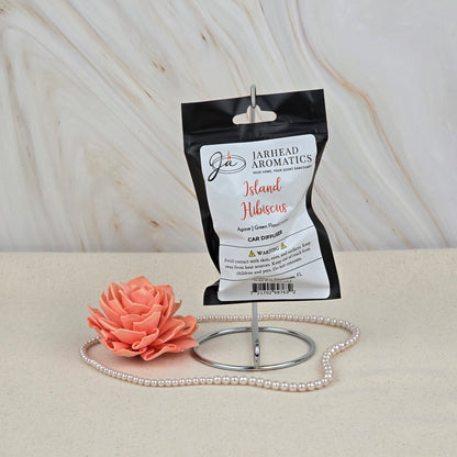The Island Hibiscus Car Diffuser by Jarhead Aromatics, a tropical floral & melon air freshener, hangs on a metal stand with a pink flower and pearls, adding refreshing fragrance on a beige surface with a marbled backdrop.
