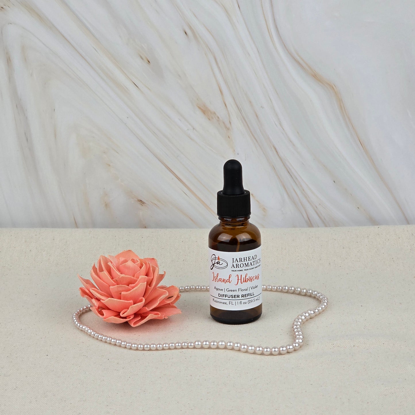 A bottle of Jarhead Aromatics Island Hibiscus Car Diffuser Refill | Tropical Floral & Melon scent rests on a beige surface next to a pink artificial flower and pearls, set against a marble-patterned background.