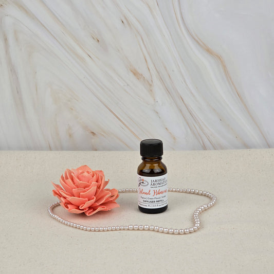 A Jarhead Aromatics Island Hibiscus Cold Air Diffuser Refill with tropical floral & melon notes, a pink artificial flower, and a string of pearls are displayed on a light surface before a marble-patterned background.