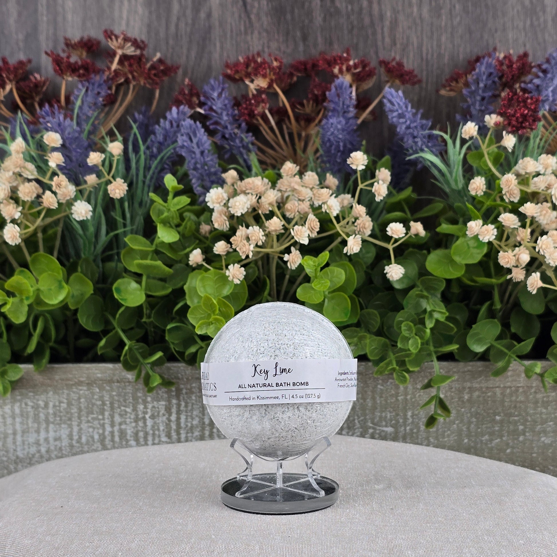 A round Key Lime Bath Bomb - All Natural by Jarhead Aromatics rests on a clear stand, framed by lush green leaves and white blossoms in a gray planter—emphasizing its pure, all-natural ingredients.