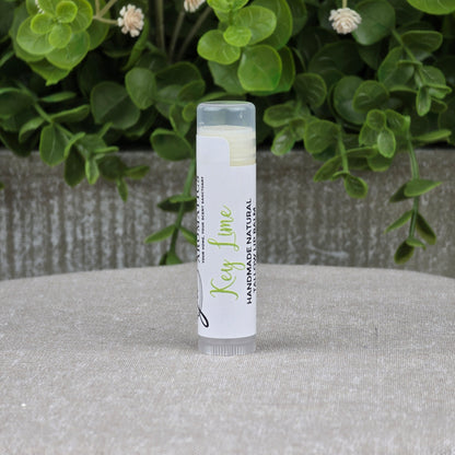 A tube of Jarhead Aromatics Tallow Handmade Natural Lip Balm - Assorted Flavors stands upright on a gray surface, framed by green leafy plants, emphasizing its natural key lime scent.