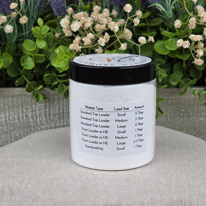 A white jar of Jarhead Aromatics Fragrance-Free Handmade Natural Laundry Soap sits on a beige surface, its label detailing machine types, load sizes, and powder amounts. Soft greenery and white flowers blur in the background.