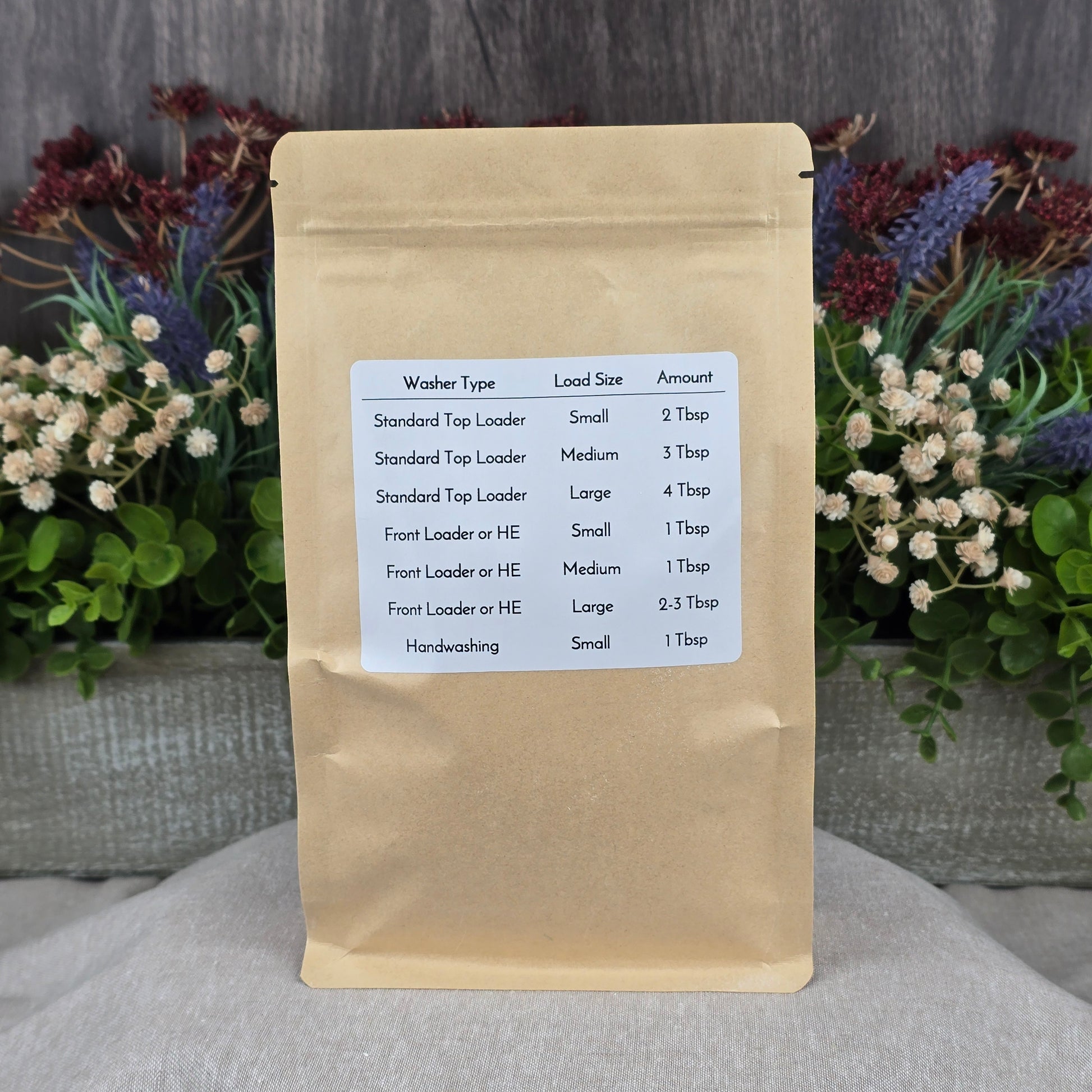 A brown paper bag of Jarhead Aromatics Fragrance-Free Handmade Natural Laundry Soap, a non-toxic powder detergent for sensitive skin, with a white label showing usage instructions and a chart for washer type, load size, and amount needed.