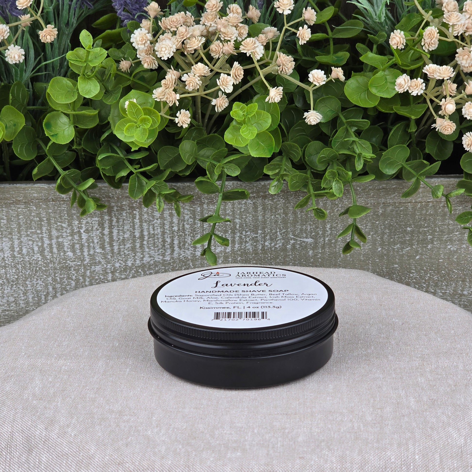 A round black container of Jarhead Aromatics Lavender Handmade Shave Soap | Beef Tallow & Goat Milk rests on a beige cushion, surrounded by green leaves and white flowers—ideal for elevating your wet shave routine.