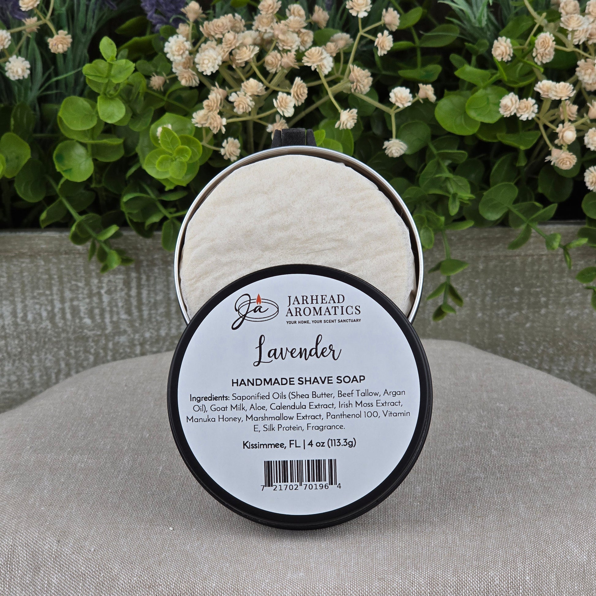 An open round container of Jarhead Aromatics Lavender Handmade Shave Soap | Beef Tallow & Goat Milk rests on a gray surface, with blurred green and purple flowers in the background—ideal for a luxurious wet shaving experience.