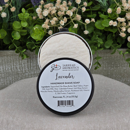 An open round container of Jarhead Aromatics Lavender Handmade Shave Soap | Beef Tallow & Goat Milk rests on a gray surface, with blurred green and purple flowers in the background—ideal for a luxurious wet shaving experience.