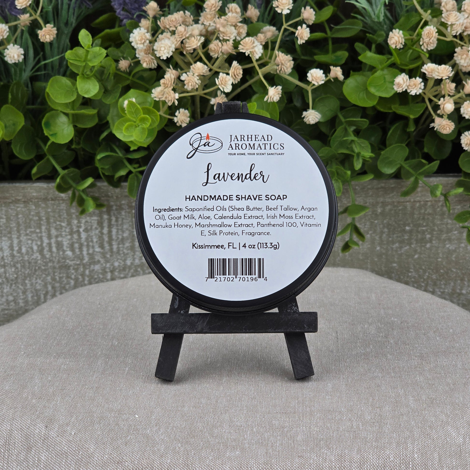 A round Jarhead Aromatics Lavender Handmade Shave Soap with beef tallow and goat milk rests on a small black easel, framed by green foliage and white flowers—an ideal choice for wet shaving enthusiasts.